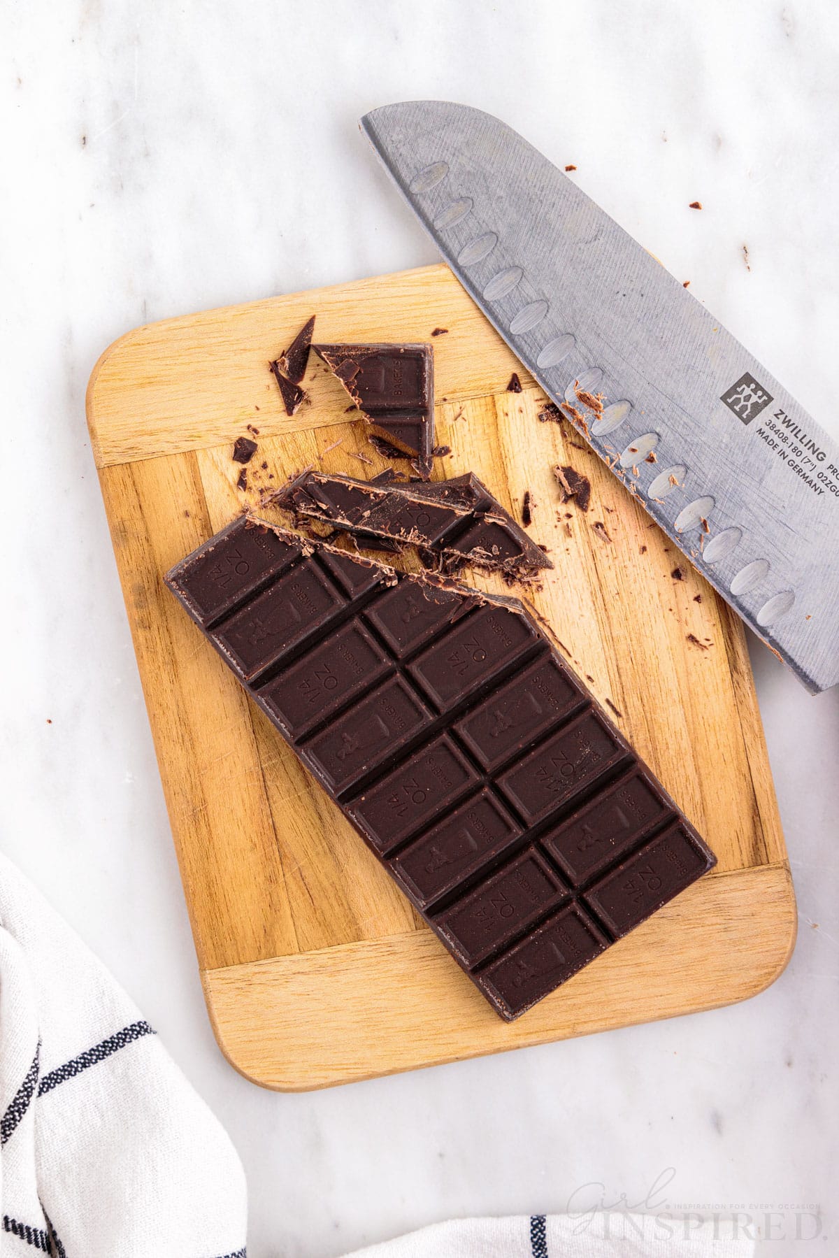 Wooden kitchen board with a chocolate bar being chopped into small pieces with a knife, checkered linen, on a marble countertop.