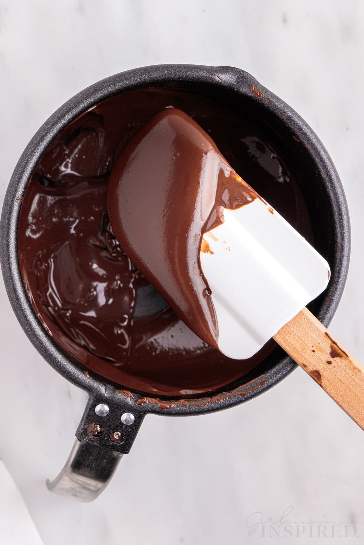 Melted chocolate in a pan with rubber spatula, on a marble countertop.