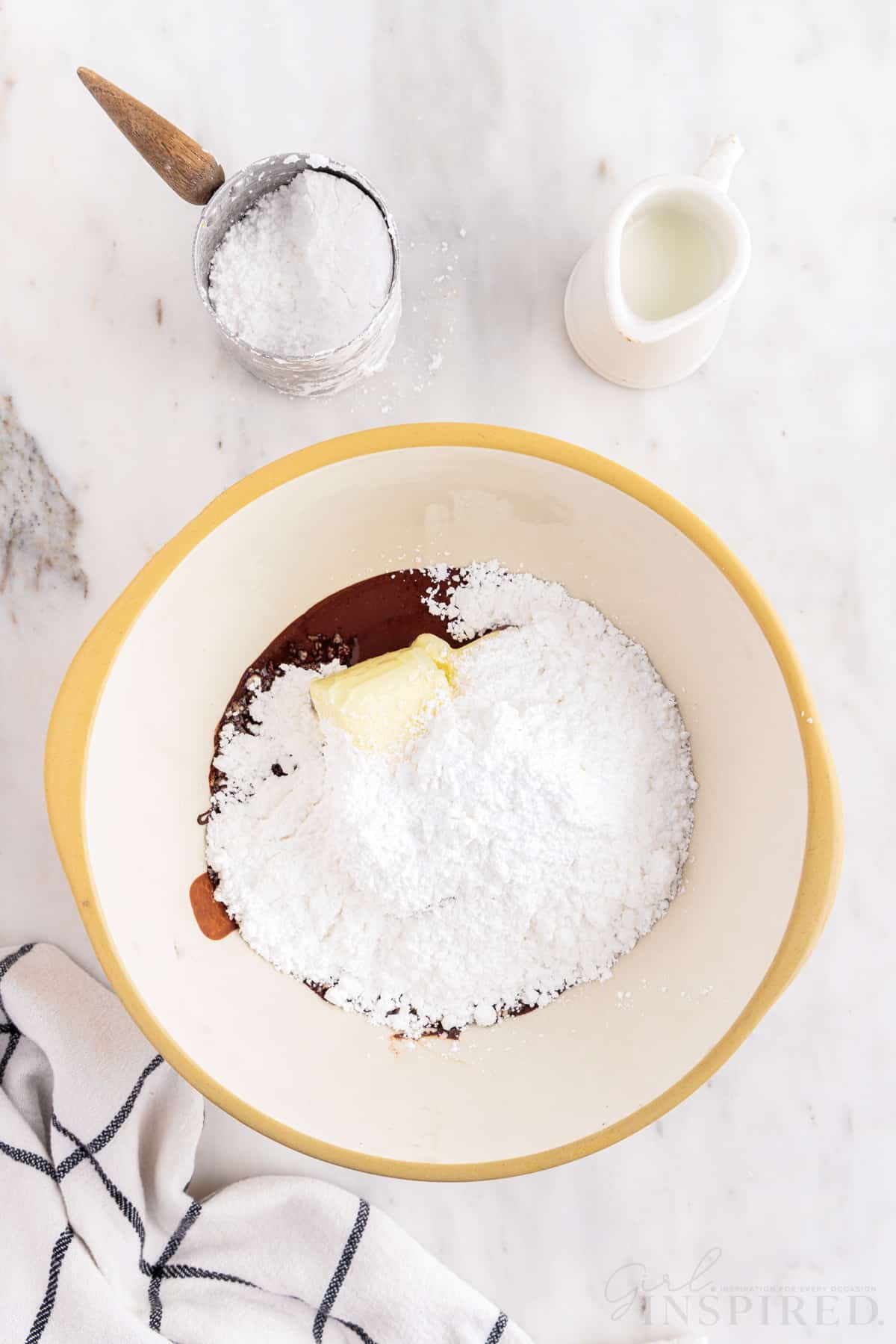 Bowl with powdered sugar, softened butter, and melted chocolate, measuring cup with powdered sugar, small jug of milk, checkered linen, on a marble countertop.