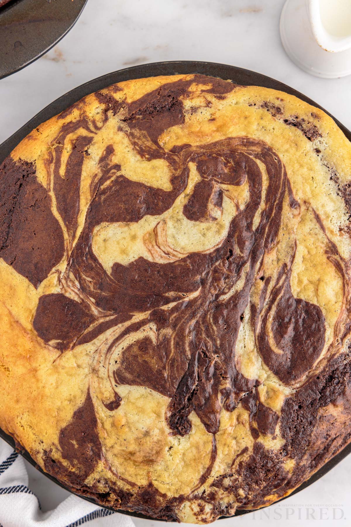 Close up of baked marble cake, checkered linen, jug of milk, on a marble countertop.