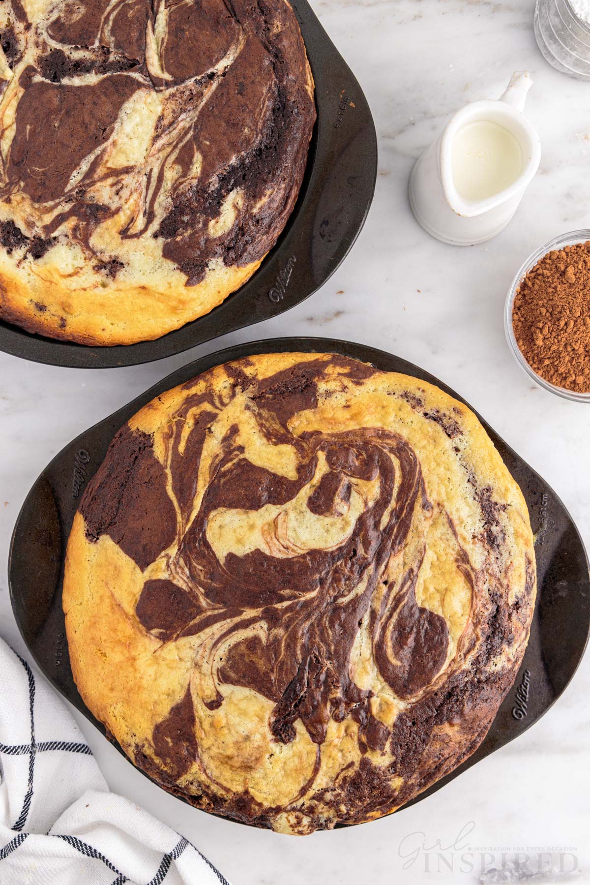 Baked marble cake in two cake pans, checkered linen, small bowl of unsweetened cocoa powder, jug of milk, on a marble countertop.