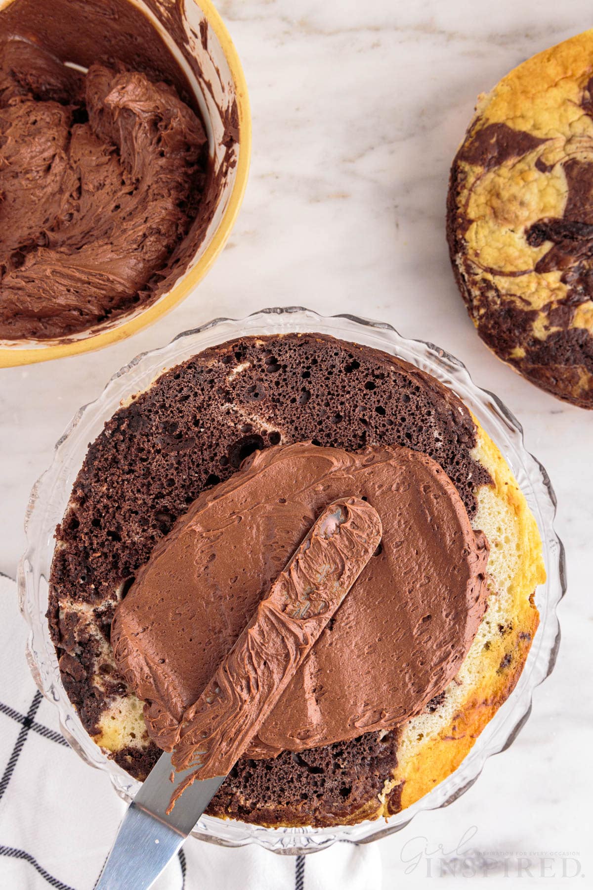 First baked marble cake layer in a glass cake dish, icing knife with chocolate frosting spread on the top of the cake, second marble cake layer, checkered linen, bowl of chocolate frosting, on a marble countertop.