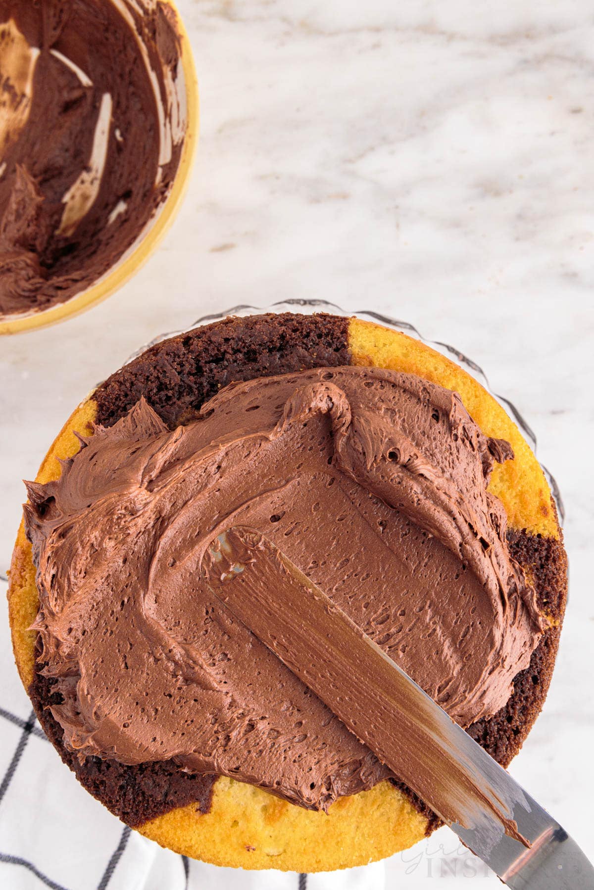 Top layer of baked marble cake being frosted with an icing knife and chocolate frosting, checkered linen, bowl of chocolate frosting, on a marble countertop.