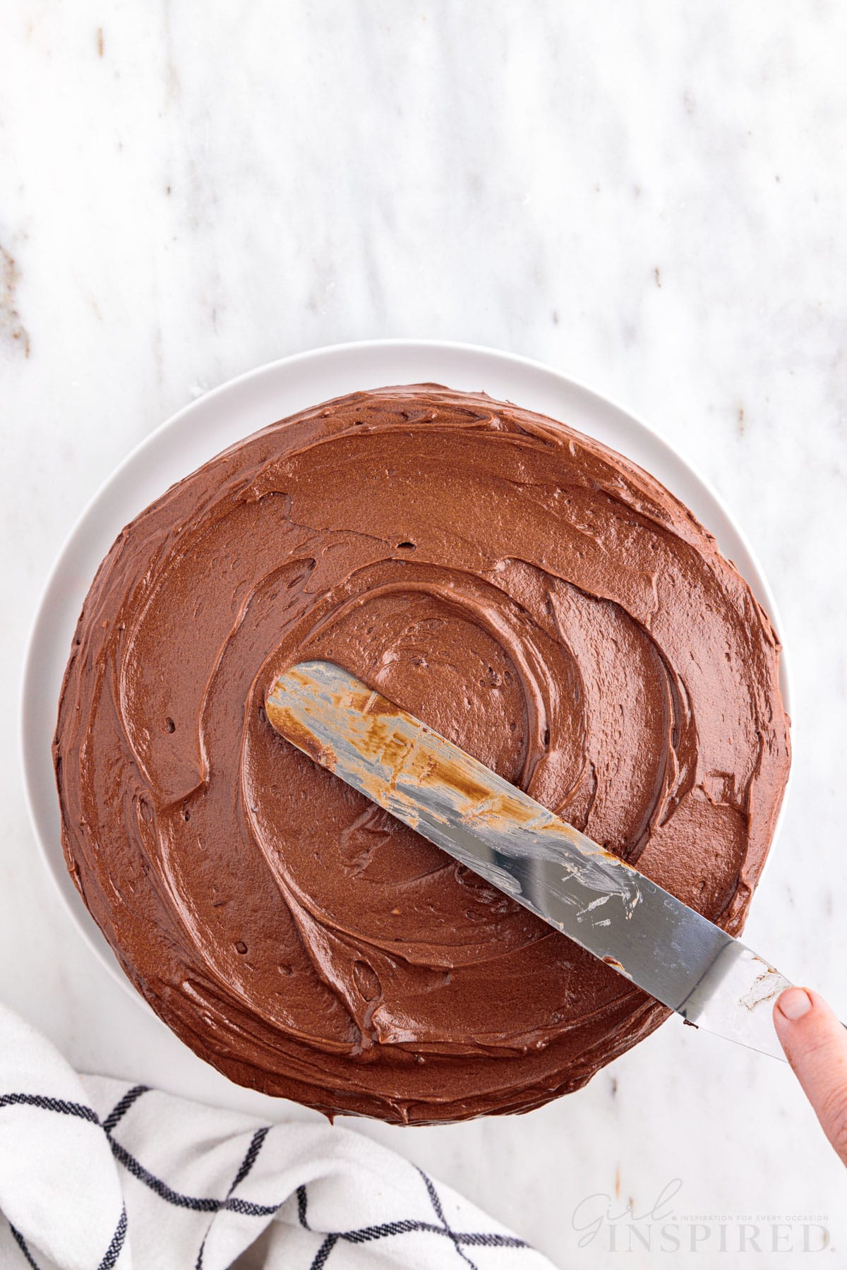 Hand smoothing chocolate frosting over the top layer of the stacked marble cake in a glass cake dish, checkered linen, on a marble countertop.
