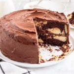 Marble cake on a large cake plate with two slices of cake removed, checkered linen, single slice of cake on a white serving plate, stacked serving plates with metal forks, jars of milk, on a marble countertop.