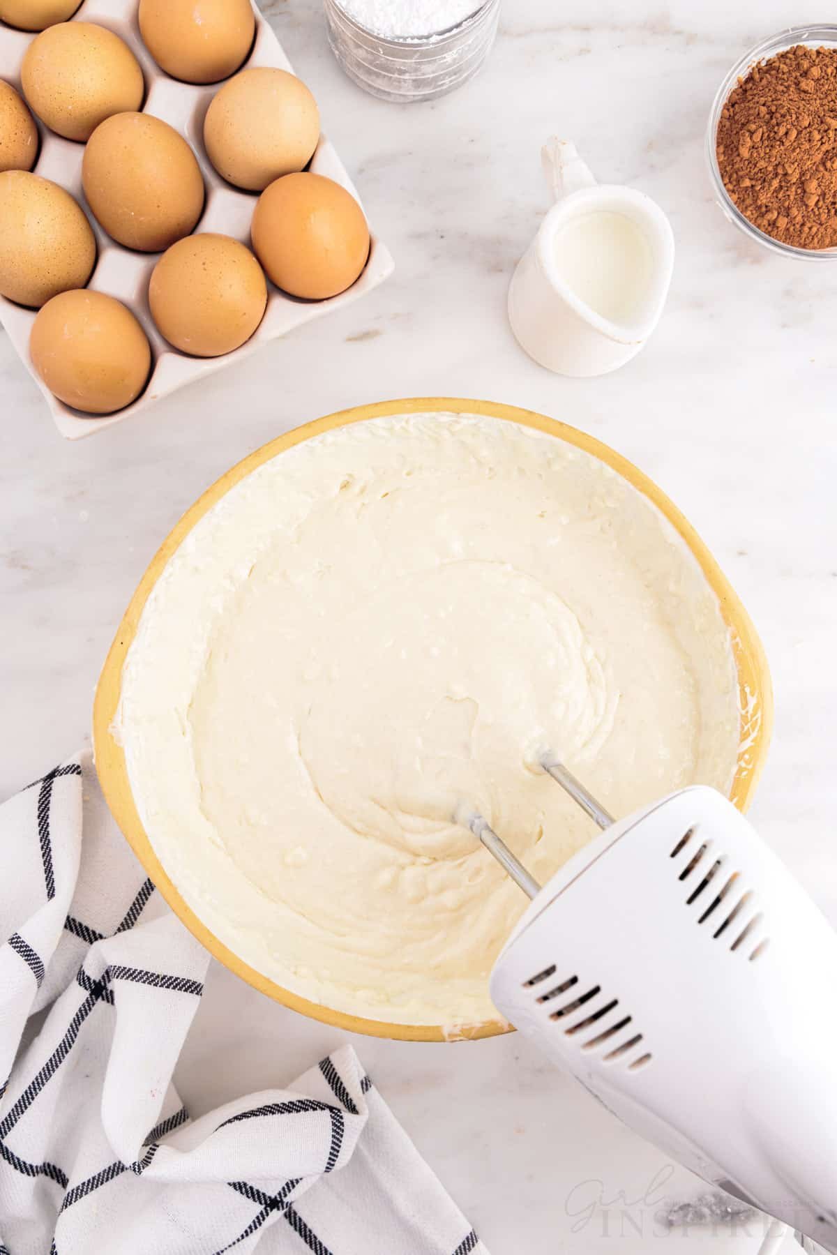Bowl with vanilla cake mix batter with electric mixer, checkered linen, tray of eggs, small jug of milk, small bowl of cocoa  powder, jug of sour cream, on a marble countertop.
