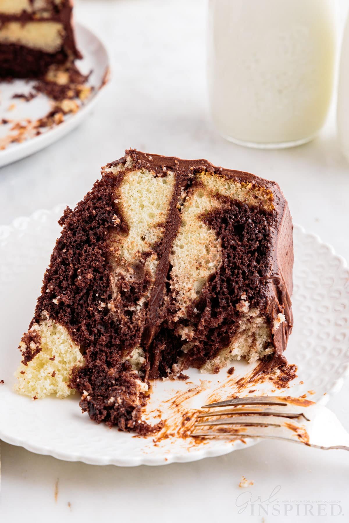 Slice of marble cake on a white serving plate with a metal fork, remaining marble cake and jug of milk in the background, on a marble countertop.