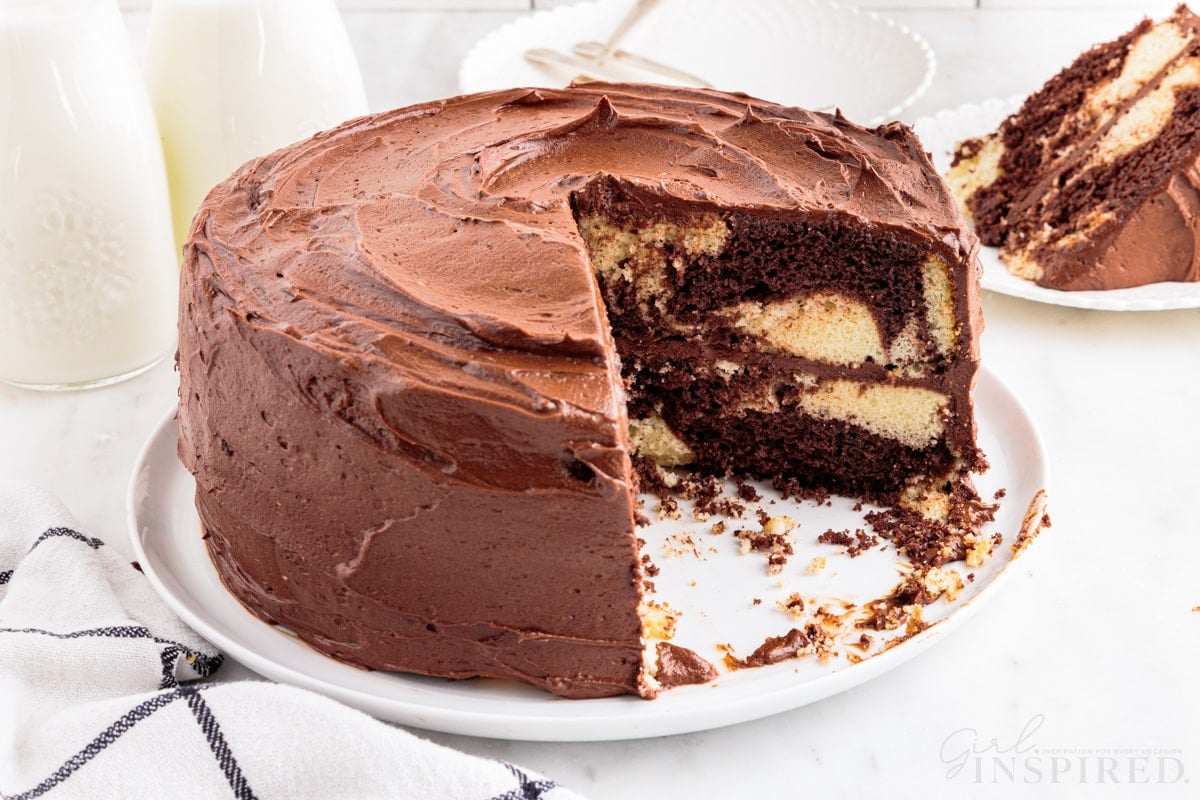 Marble cake on a large serving plate with two slices of cake removed, one slice of marble cake on a white side plate, checkered linen, two jars of milk, on a marble countertop.