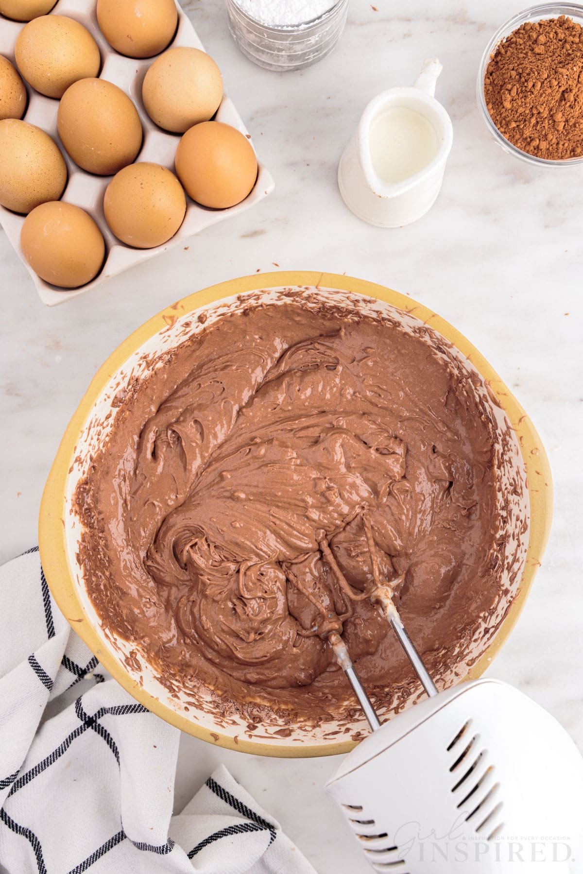 Bowl of chocolate cake mix batter with electric beater, tray of eggs, checkered linen, small bowl of cocoa powder, small jug of milk, jug of sour cream, on a marble countertop.