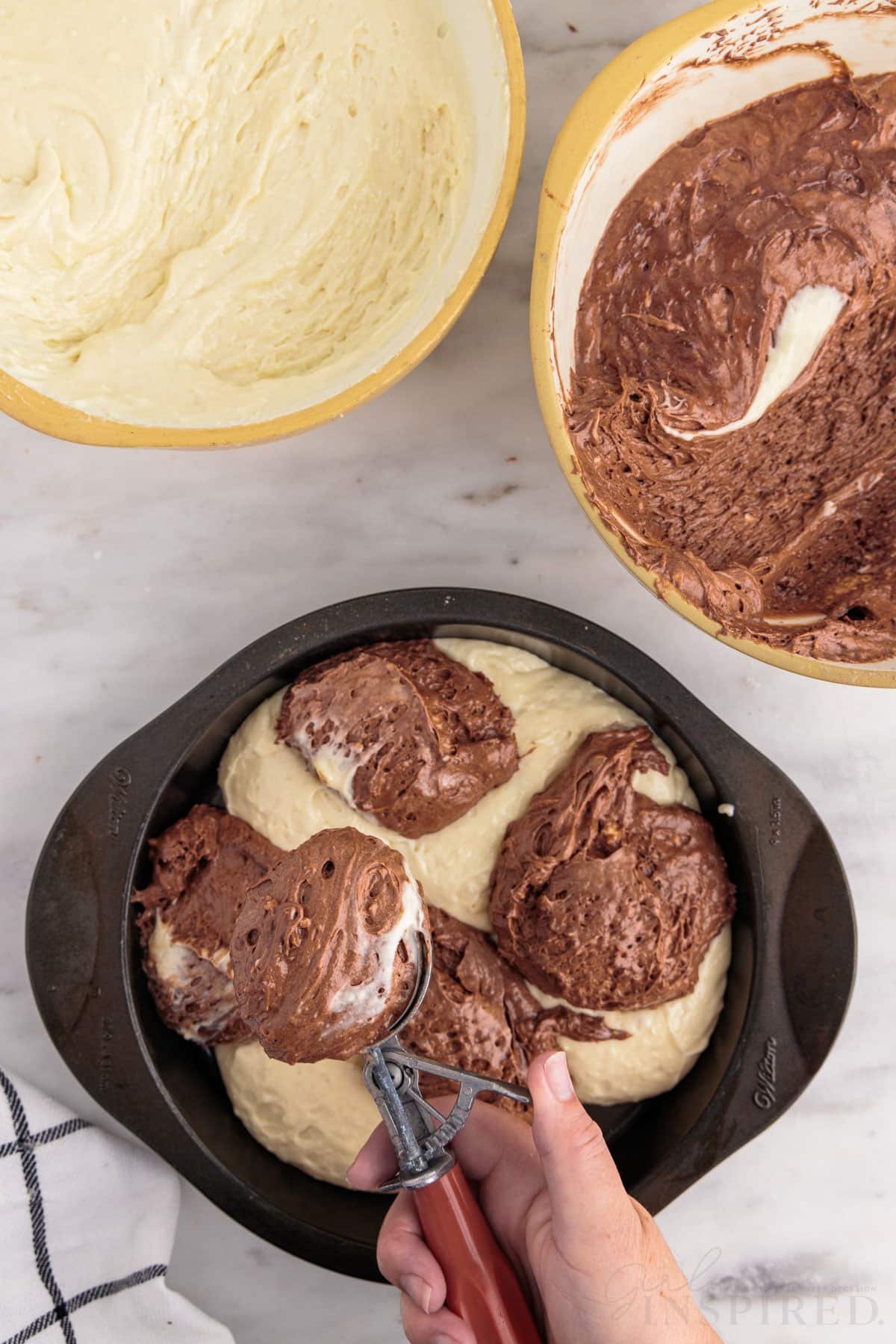 Bowl of vanilla cake batter, bowl of chocolate cake batter, prepared cake pan with hand over dropping scoops of chocolate cake mix into the pan, checkered linen, on a marble countertop.