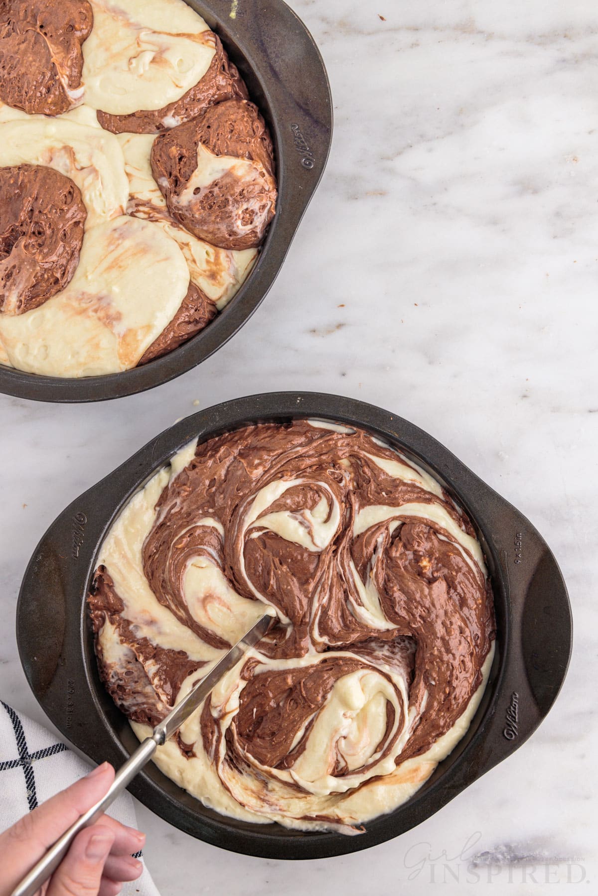 Cake pan with alternating scoops of vanilla and chocolate cake mix batter, second cake pan with vanilla and chocolate cake mix and hand over the mix with a metal knife gently creating swirls in the batter, checkered linen, on a marble countertop.