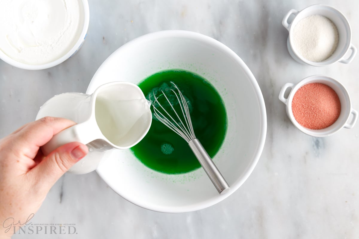 water poured into a mixing bowl to make green jello with whisk next to jello and sugar