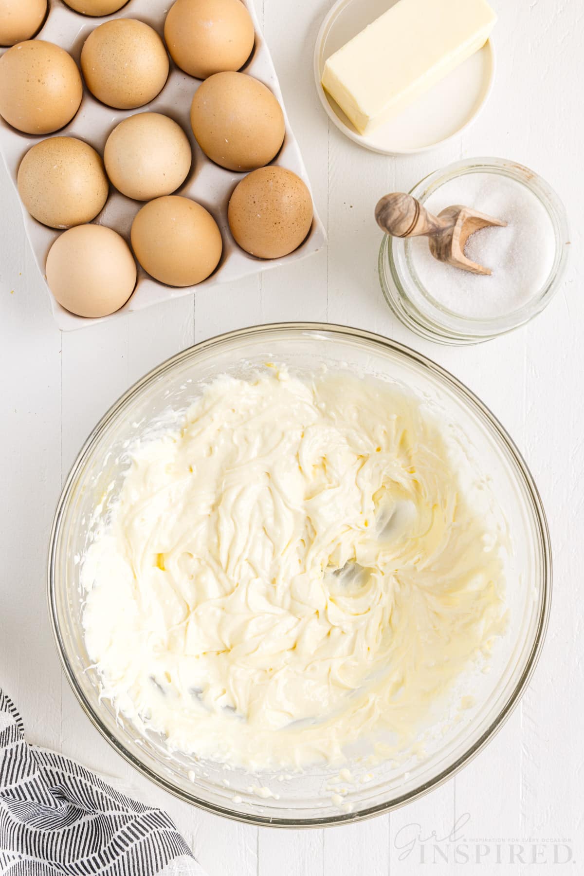 Cream cheese mixture in a glass bowl, tray of eggs, stick of butter, jar of sugar, textile linen, on a marble countertop.