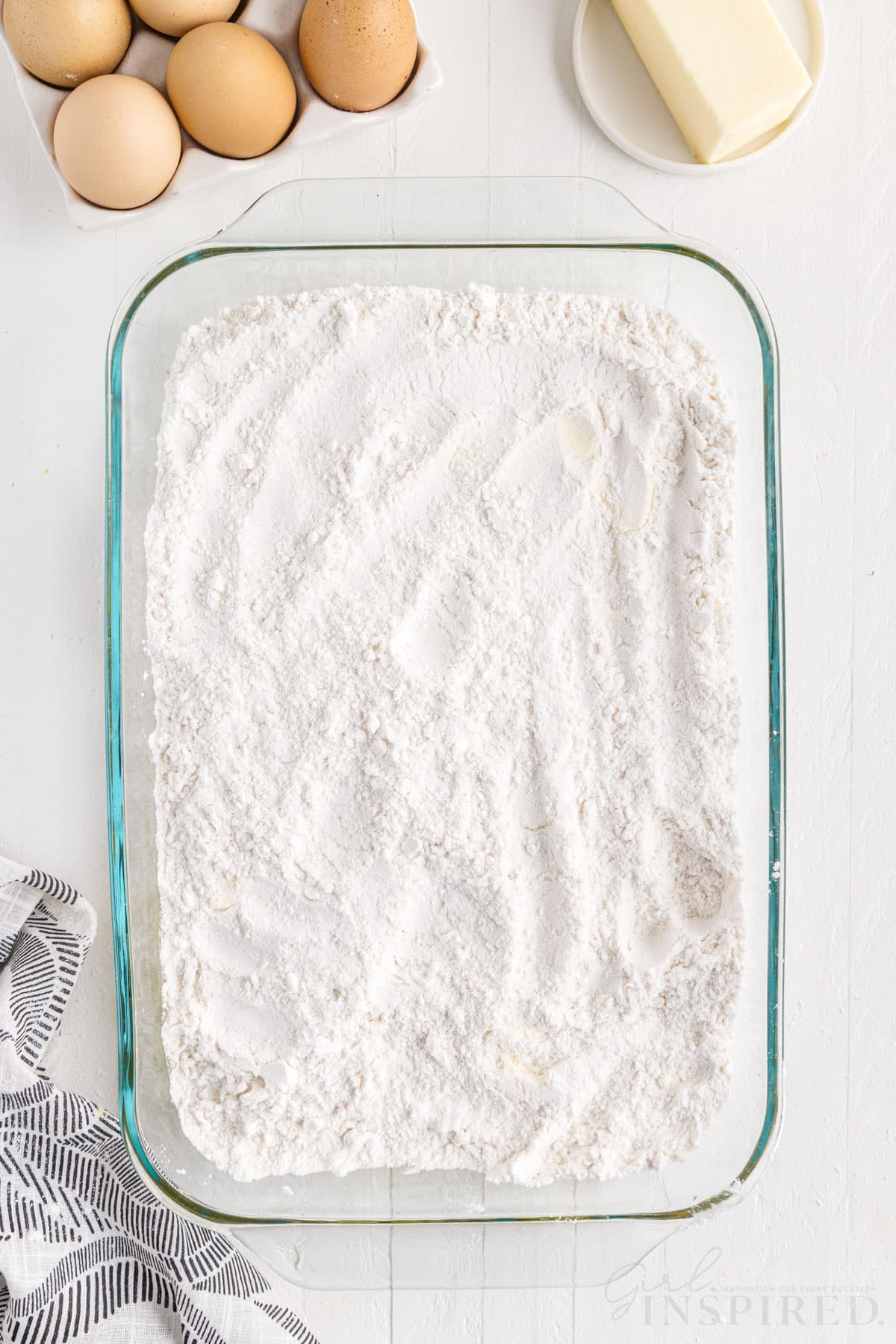 Baking dish with white cake mix on top of the cream cheese mixture, tray of eggs, stick of butter, textile linen, on a marble countertop.
