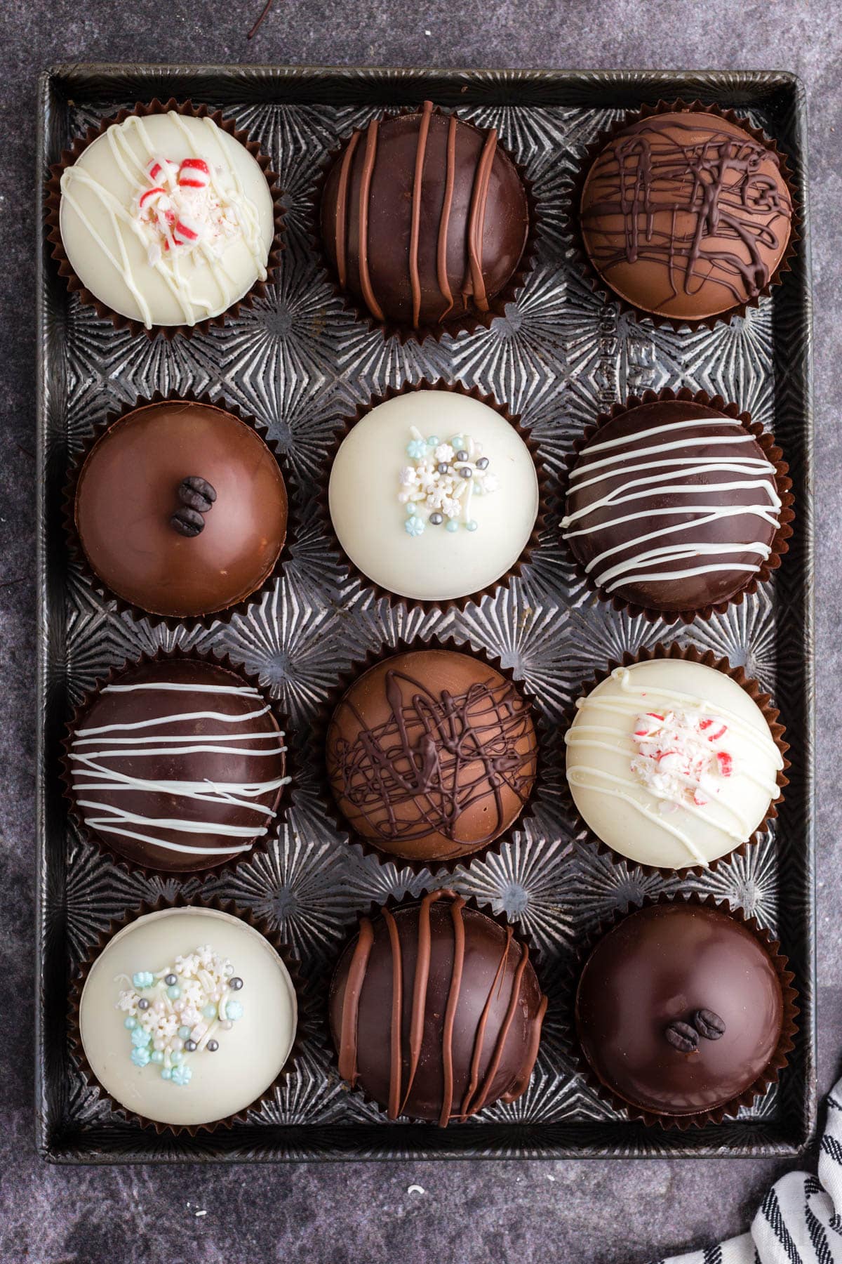 Assorted hot chocolate bombs on a metal tray, striped linen, on a dark marble countertop.
