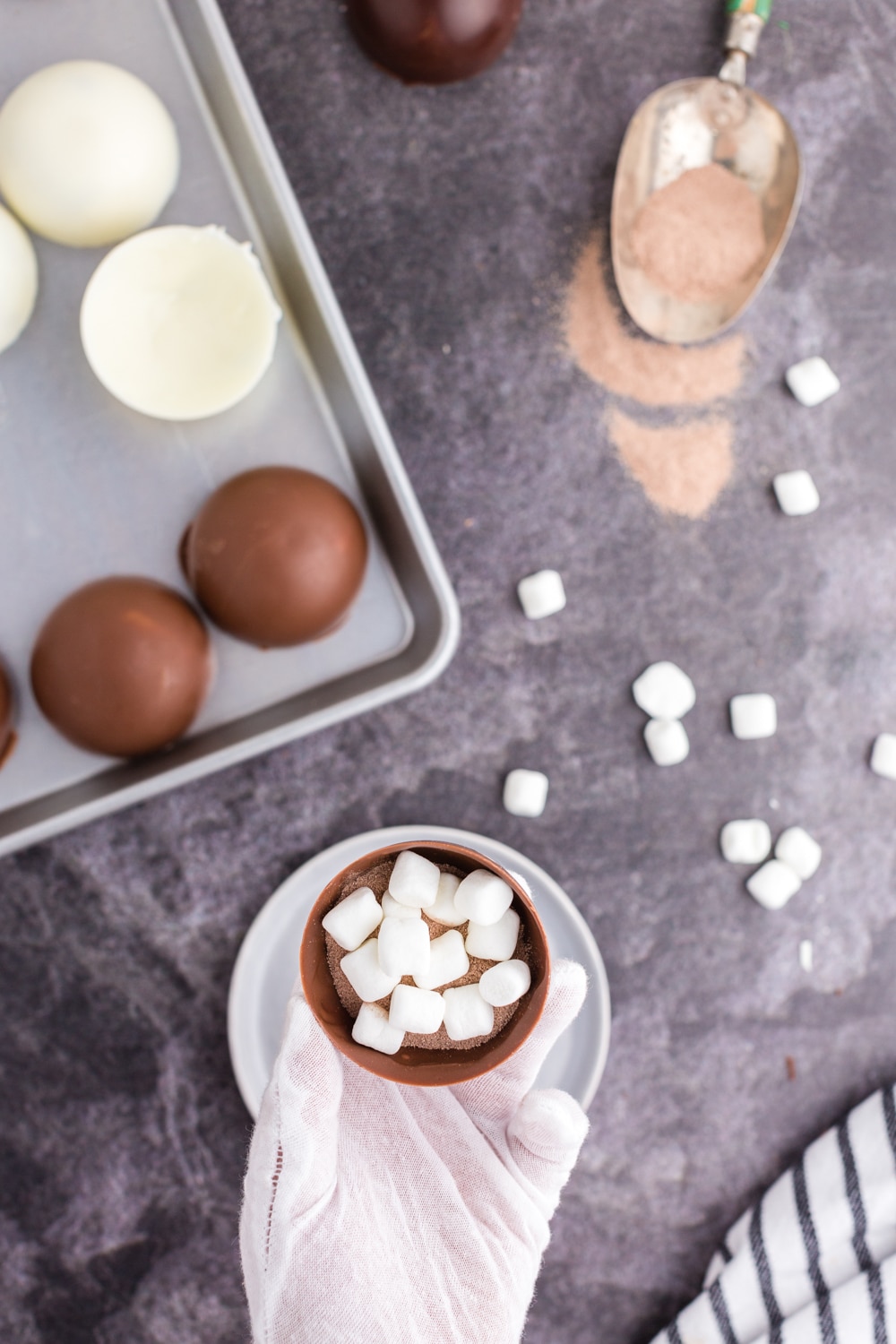 Hand with glove holding one sphere that's filled with hot chocolate and mini marshmallows, tray of chocolate spheres, metal scoop with hot chocolate, loose mini marshmallows, striped linen on a dark marble countertop.