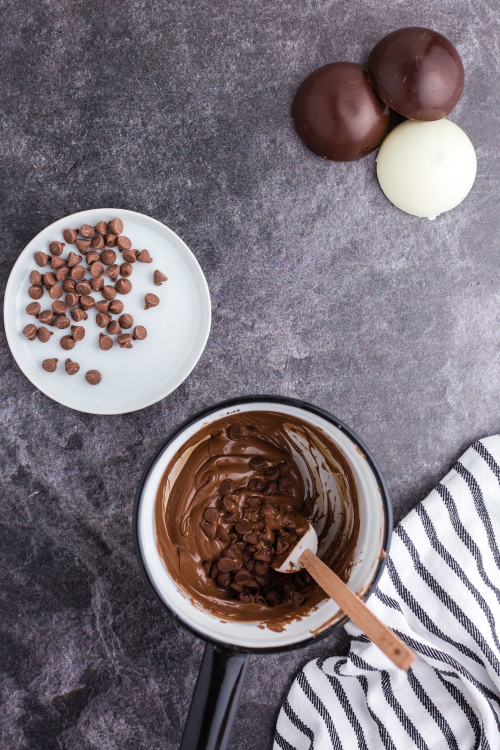 Bowl with melted chocolate chips and small spatula, shallow dish with remaining chocolate dips, striped linen, hot chocolate bomb halves, on a dark marble countertop.