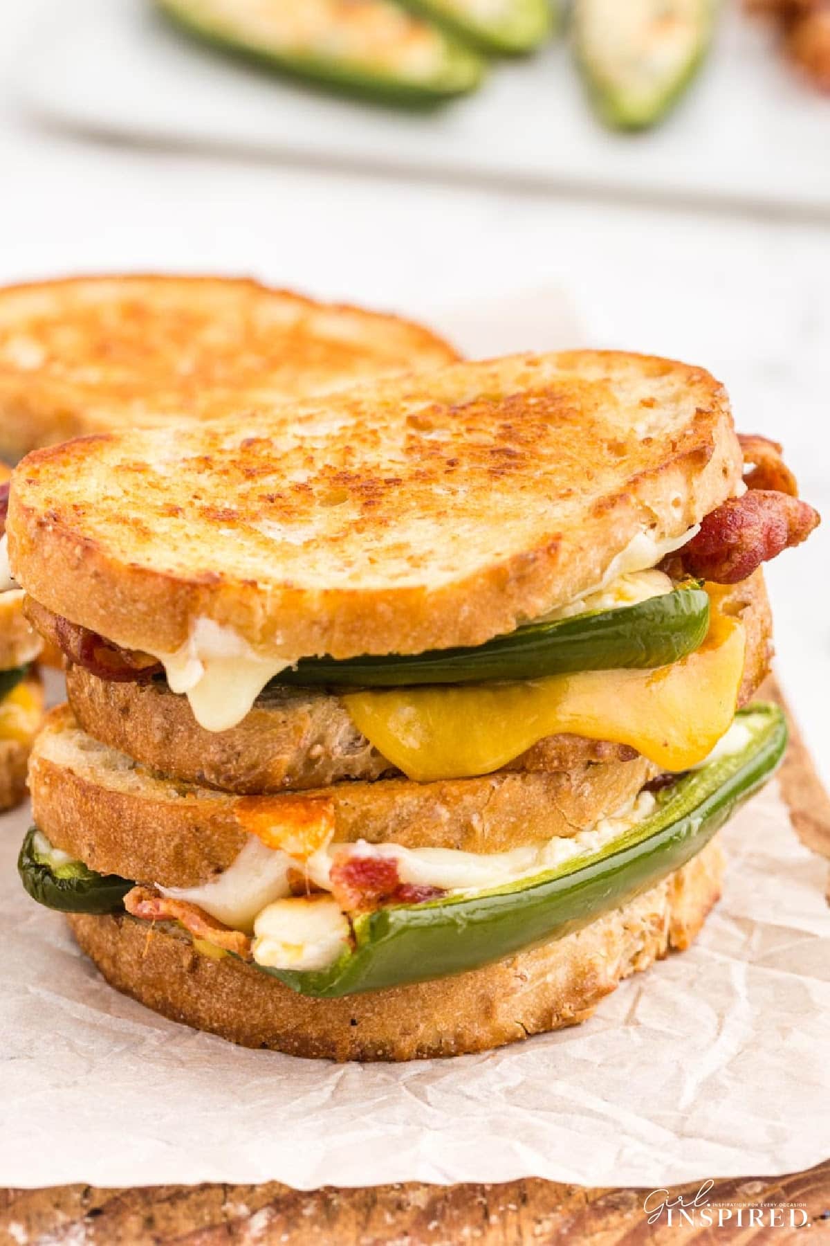 Two jalapeño popper grilled cheese sandwiches stacked on parchment paper-lined wooden kitchen board, stuffed jalapeno poppers, crispy bacon strips, on a white marble countertop.