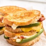 Two Jalapeño popper grilled cheese sandwiches stacked on parchment paper-lined wooden kitchen board, stuffed jalapeno poppers, crispy bacon strips, on a white marble countertop.
