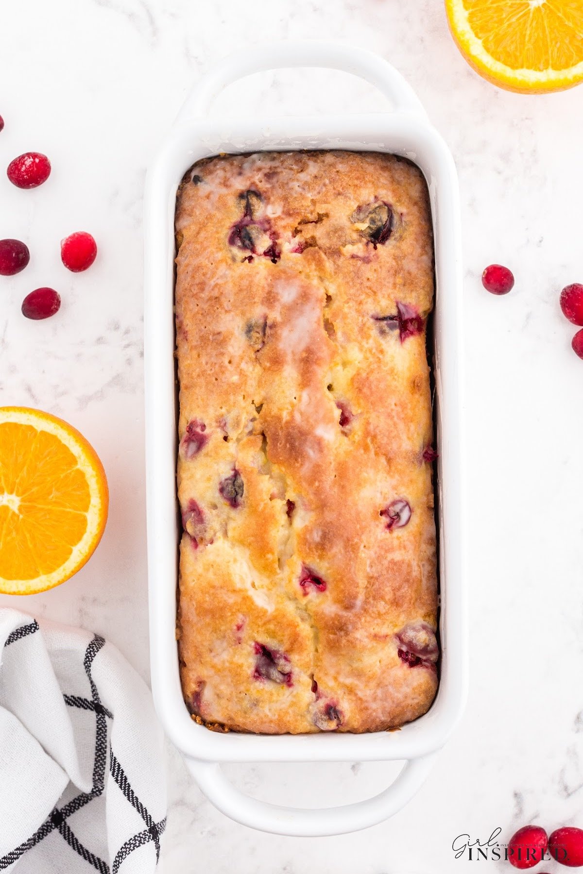 Glazed orange cranberry bread in loaf pan, with fresh oranges and cranberries scattered around.