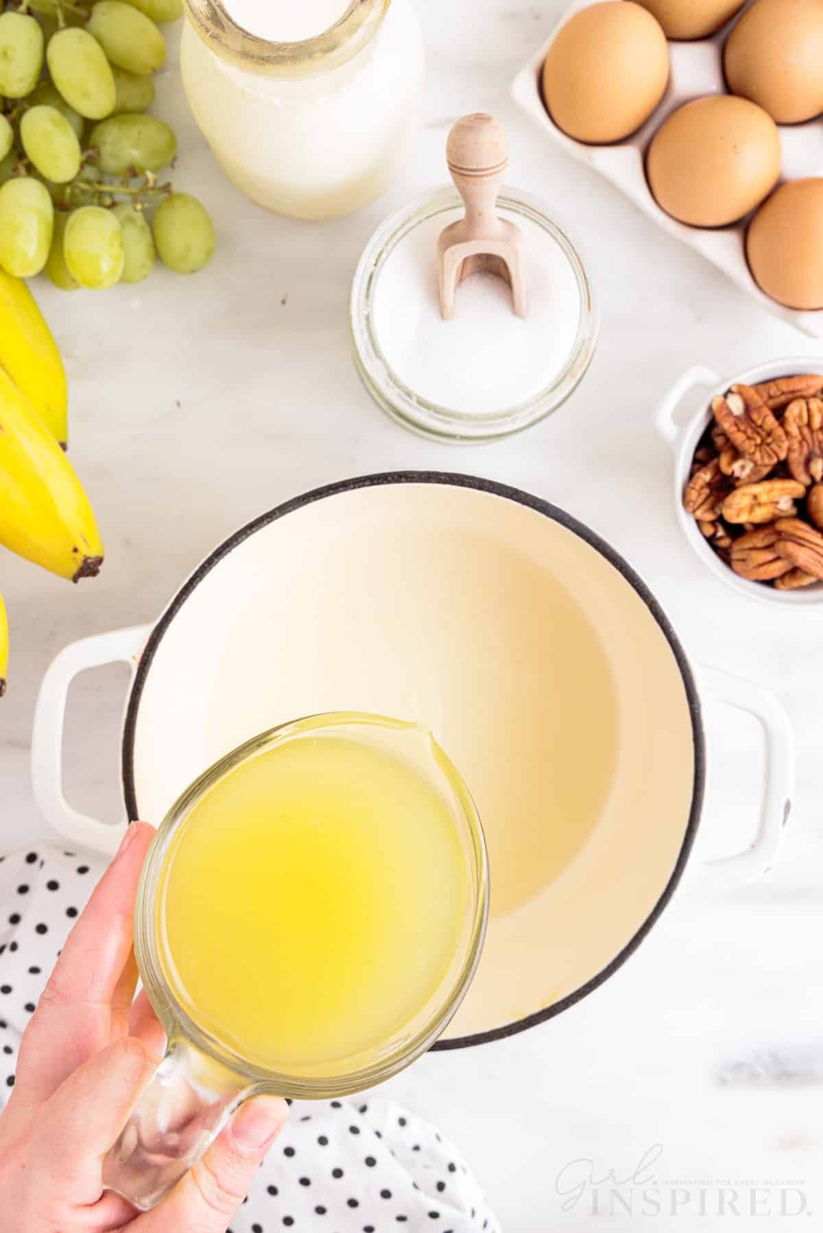 Hand pouring pineapple and orange juices into a saucepan, bunch of bananas, polka dot linen, green grapes, bowl of granulated sugar, tray of eggs, bowl of pecans, jar of heavy cream, on a marble countertop.