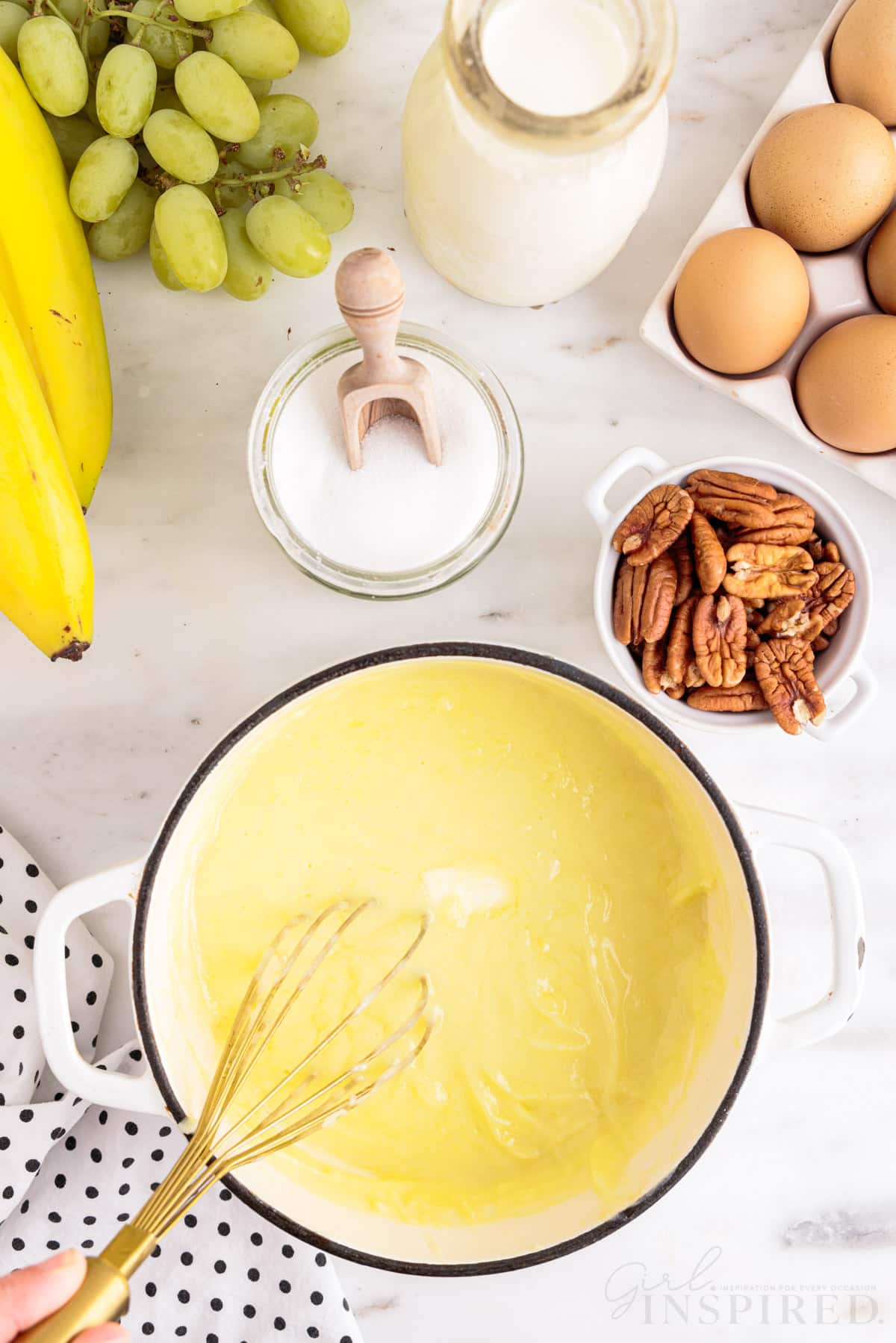 Saucepan with thickened custard, hand holding metal whisk in the custard mixture, polka dot linen, bunch of bananas, green grapes, bowl of granulated sugar, tray of eggs, bowl of pecans, jug of heavy cream, on a marble countertop.