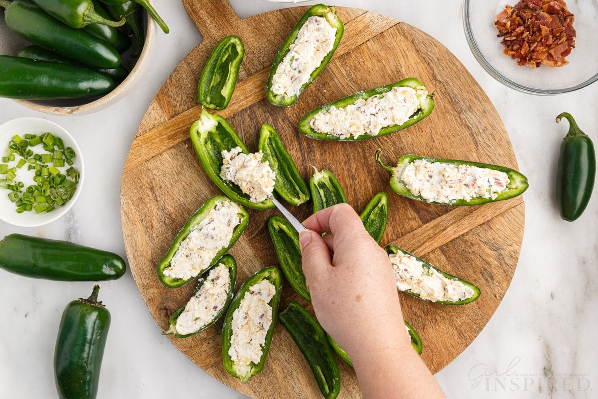jalapenos stuffed with cream cheese mixture on wooden try next to jalapenos and bacon.