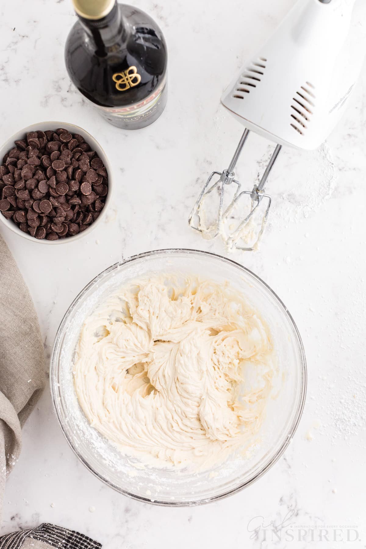 Bowl of buttercream, electric mixer, bottle of Bailey's Irish cream, bowl of chocolate chips, linen, on a marble countertop.