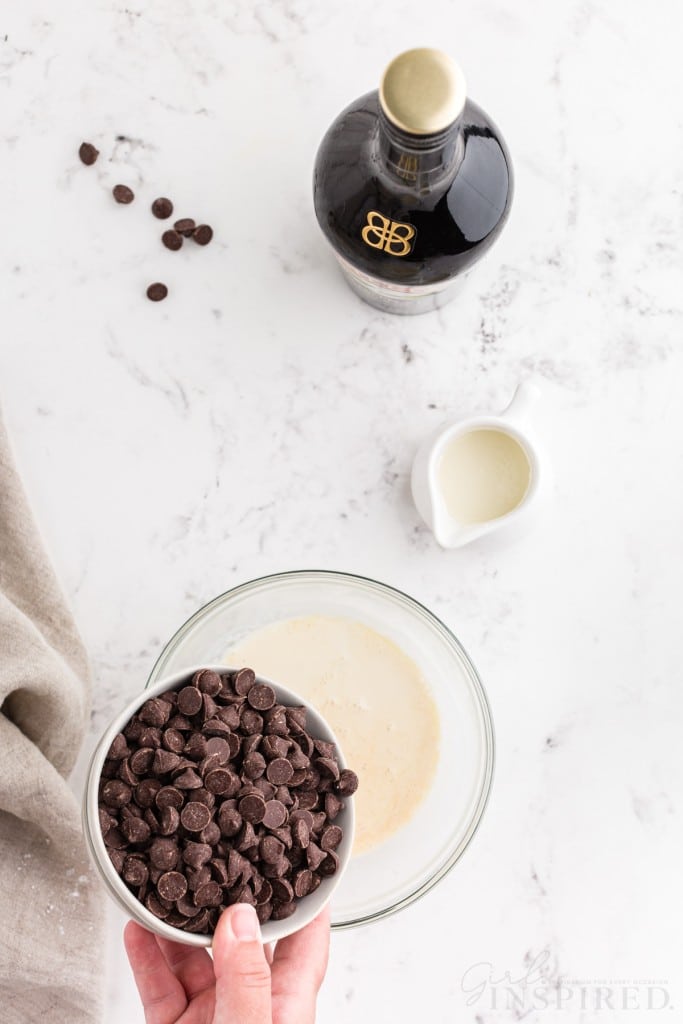 Hand pouring chocolate chips into the bowl with heavy whipping cream and Bailey's Irish cream, jug of heavy whipping cream, linen, bottle of Bailey's Irish cream, on a marble countertop.