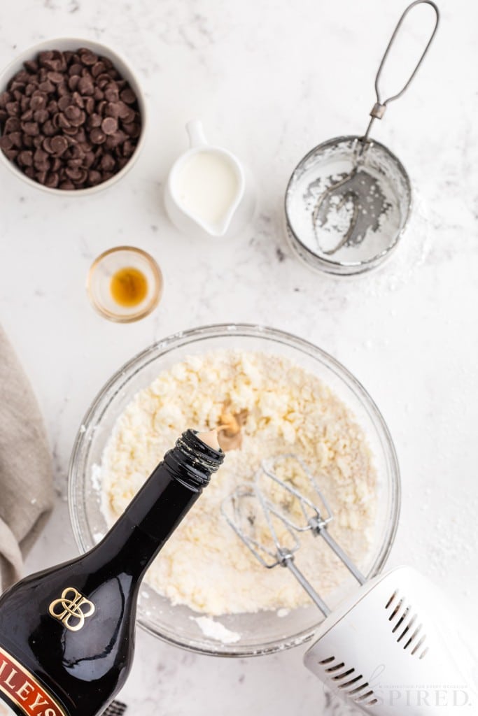 Bowl with beaten butter and sugar mixture, electric mixer with attachments in the bowl, bottle of Bailey's Irish cream poured into the bowl, linen, bowl of chocolate chips, jug of heavy whipping cream, on a marble countertop.