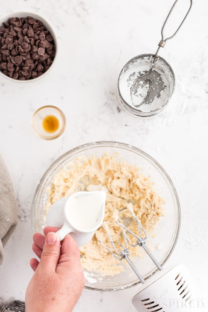 Mixing bowl of buttercream mixture with hand pouring heavy whipping cream into the bowl, electric mixer with attachments in the bowl, linen, chocolate chips, empty measuring cup, on a marble countertop.