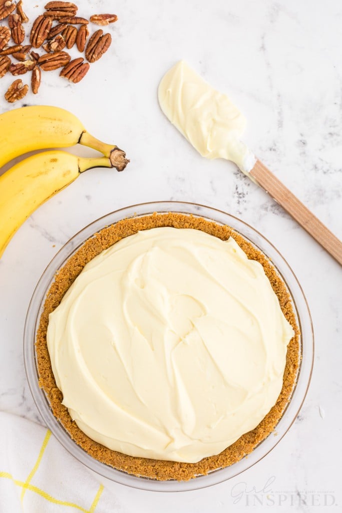 Banana cream pie without any garnish, whole bananas, plastic spatula with pudding filling, pecans, on a marble countertop.