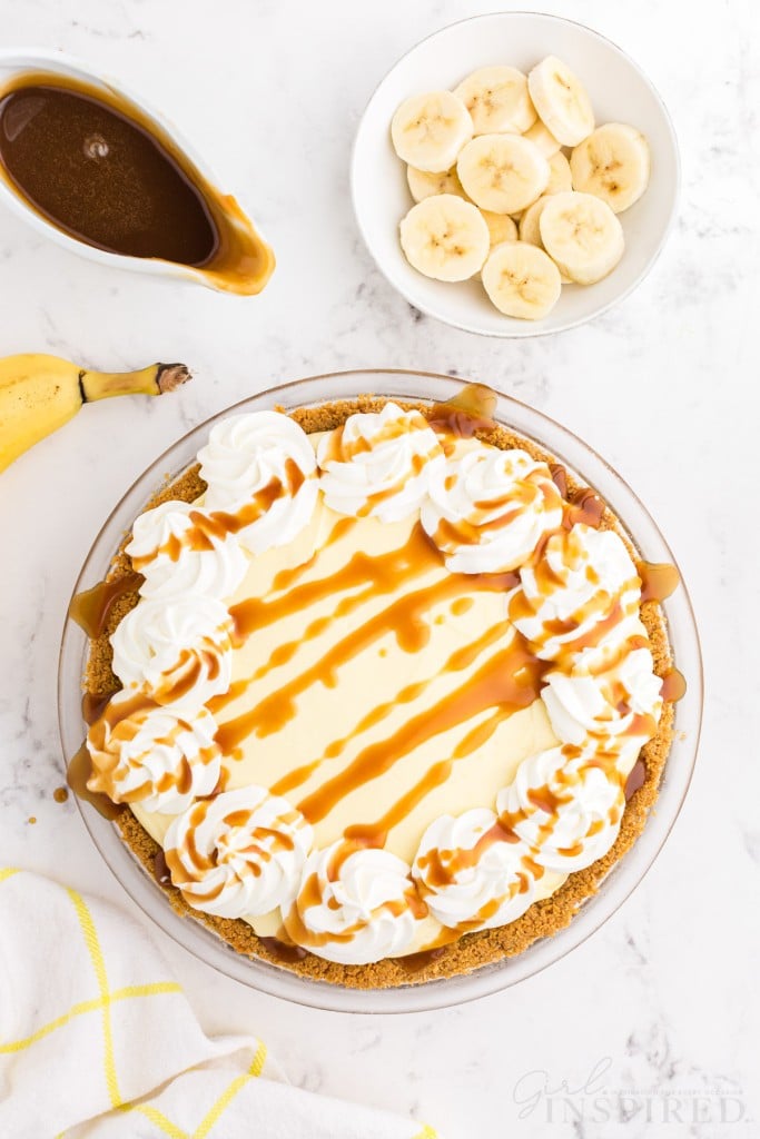 Banana cream pie with Graham cracker crust topped with piped whipped cream and caramel drizzle, bowl of sliced bananas, jug of caramel, whole banana, yellow striped linen, on a marble countertop.