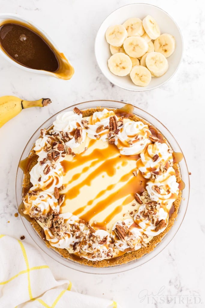 Banana cream pie with Graham cracker crust topped with whipped cream, caramel, and chopped pecans, whole banana, jug of caramel, bowl of sliced banana, yellow striped linen, on a marble countertop.