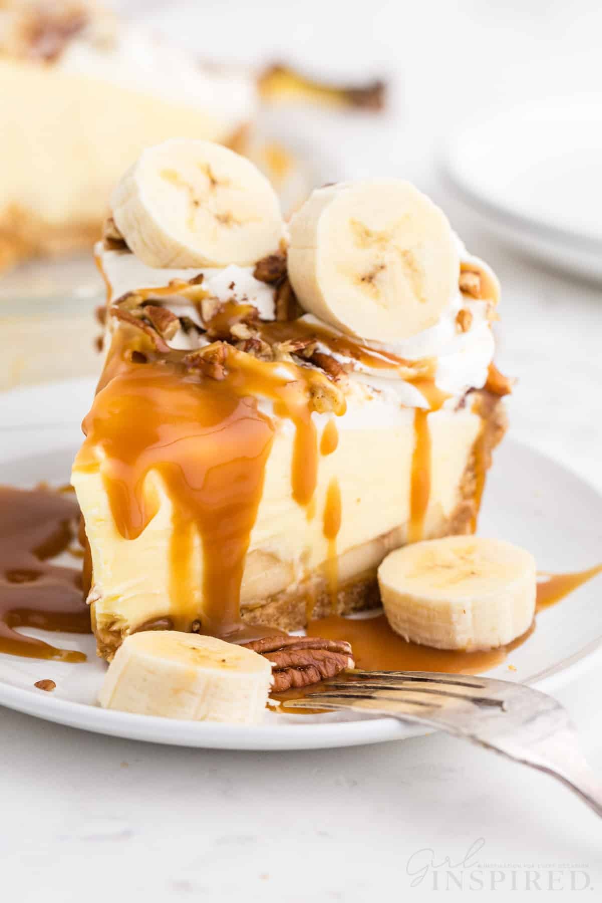 Banana cream pie with Graham cracker crust on a white serving plate with a metal fork, banana cream pie in glass baking pie dish in the background, stacked white plates, on a marble countertop.