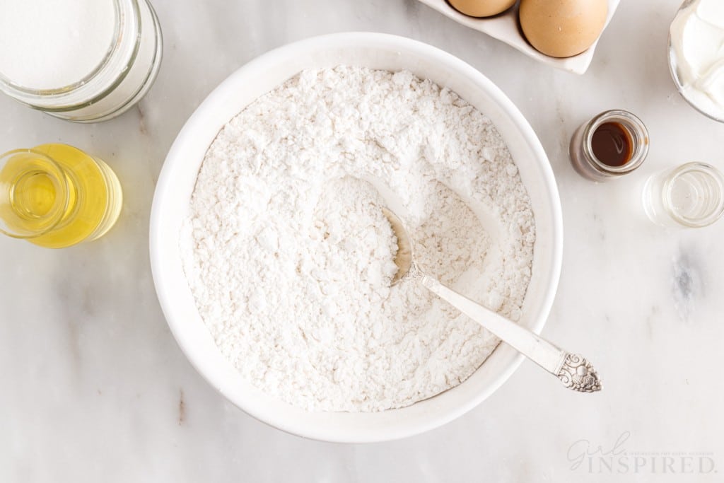 flour in bowl with spoon next to oil, vanilla, and eggs