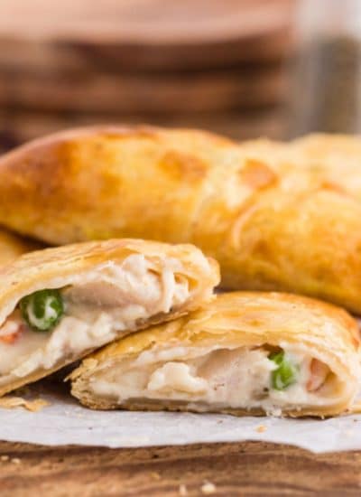 Baked chicken pot pie hand pies stacked on top a wooden board with parchment paper, a pie cut in half with the filling coming out