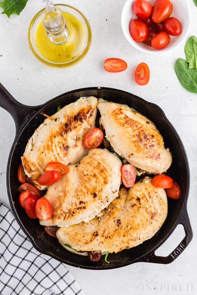 Four browned chicken breasts in a skillet with halved grape tomatoes, olive oil, bowl of grape tomatoes, baby spinach, checkered linen, on a marble countertop.