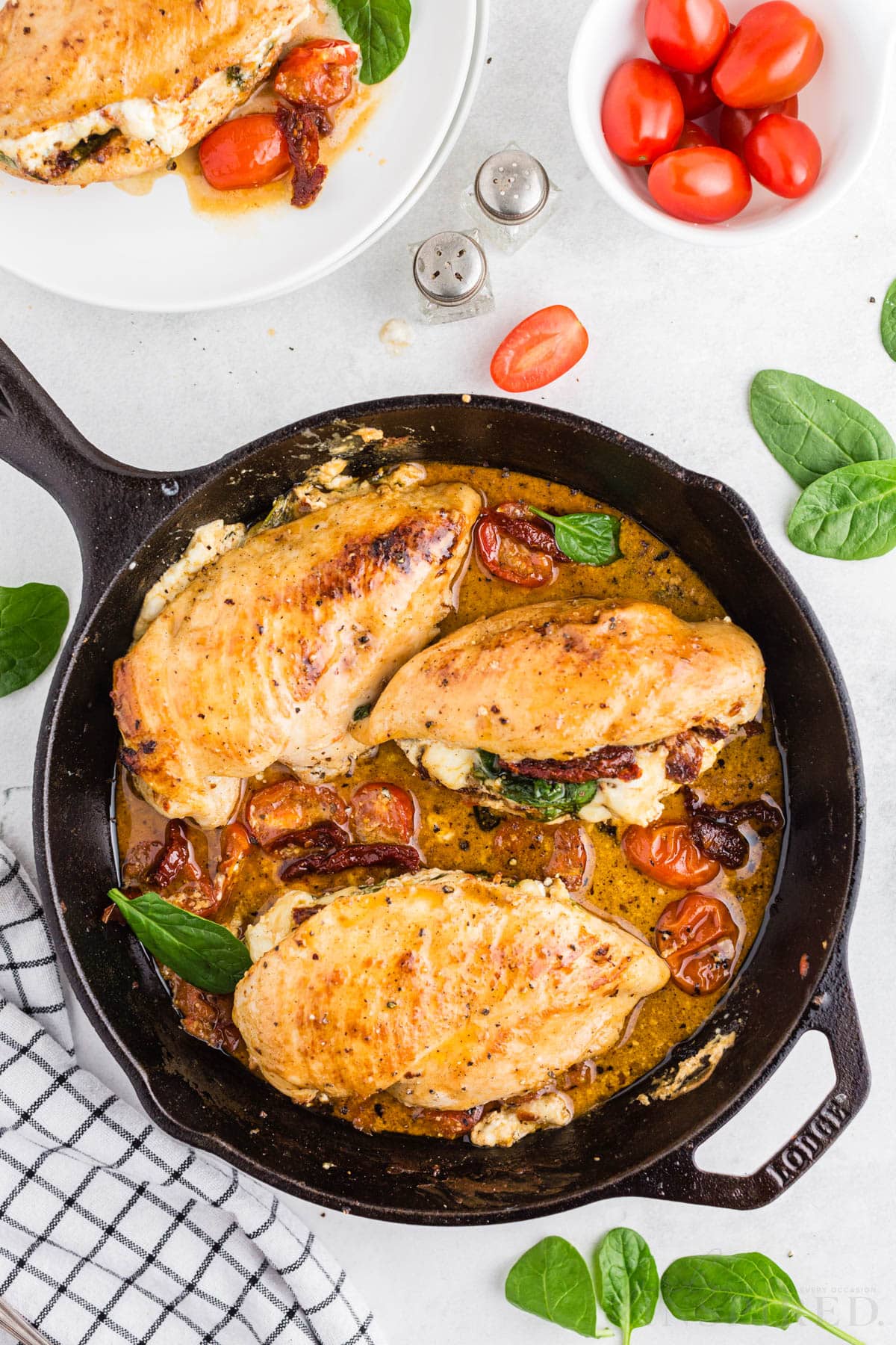 Chicken with sundried tomatoes and spinach in a cast iron skillet, checkered linen, plate with stuffed chicken breast, small bowl of tomatoes, salt and pepper shakers, on a marble countertop.