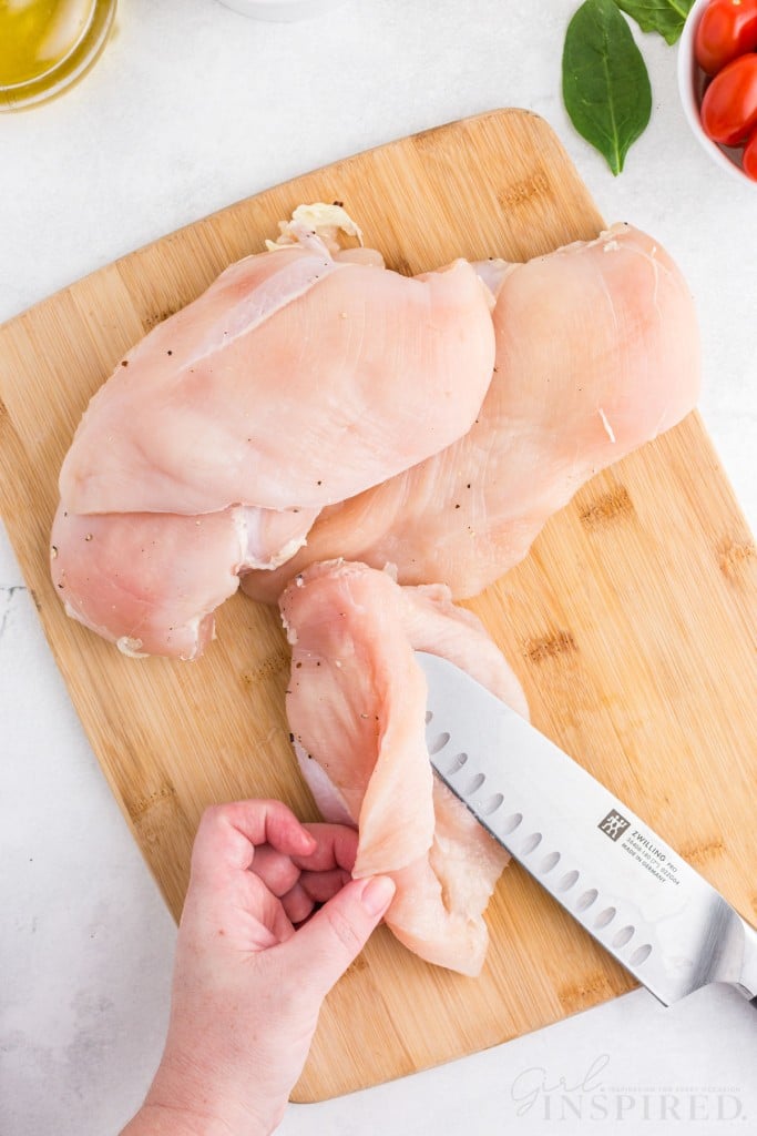 Hand holding a chicken breast while slicing the chicken breast along the side with a sharp kitchen knife, chicken breasts on a wooden kitchen board, on a marble countertop.