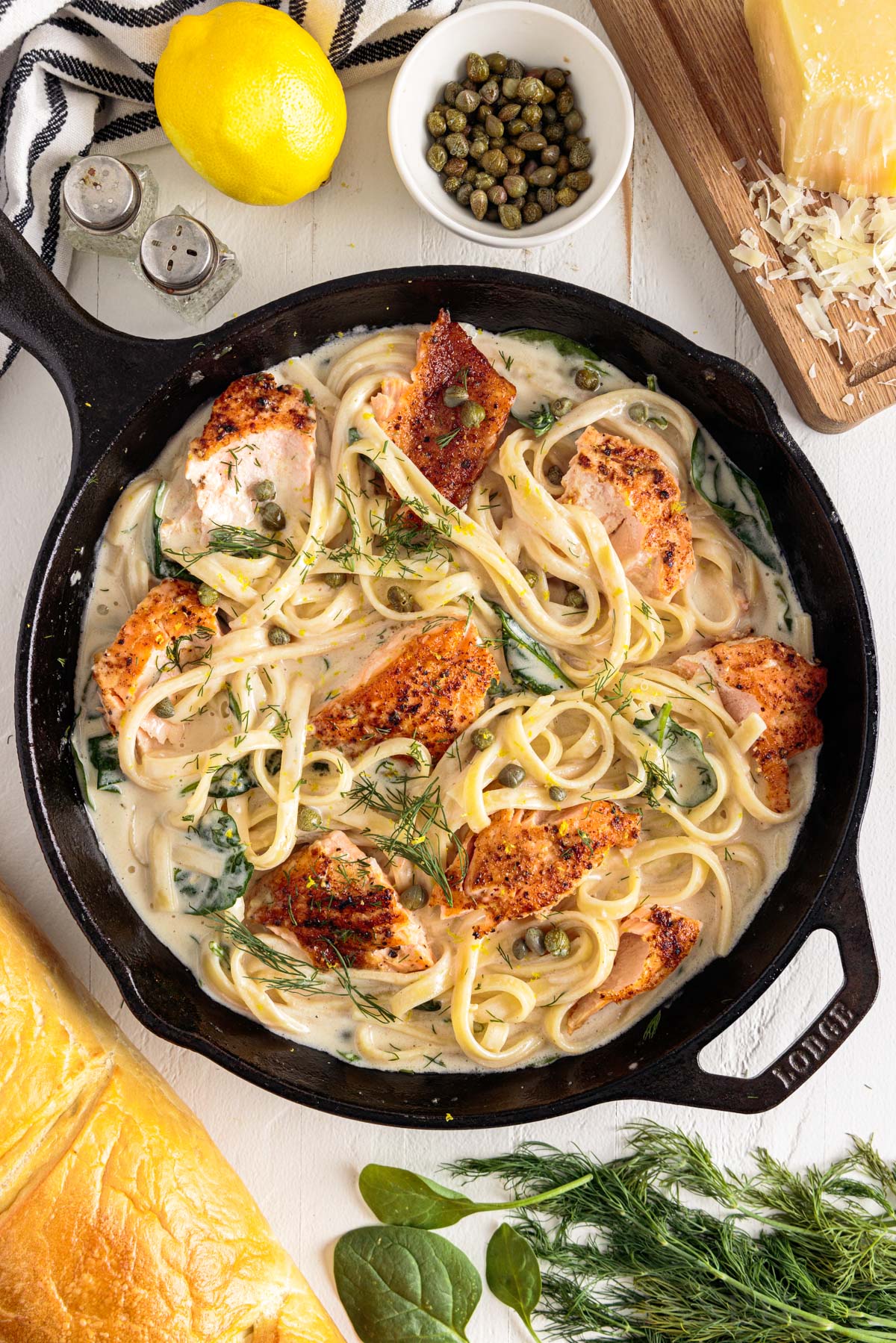 top full view of creamy salmon pasta in skillet next to lemon, capers, salt and pepper, cheese, spices, and bread