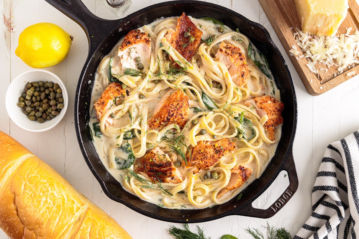 top view of creamy salmon pasta in a skillet next to bread, capers, lemon, and cheese