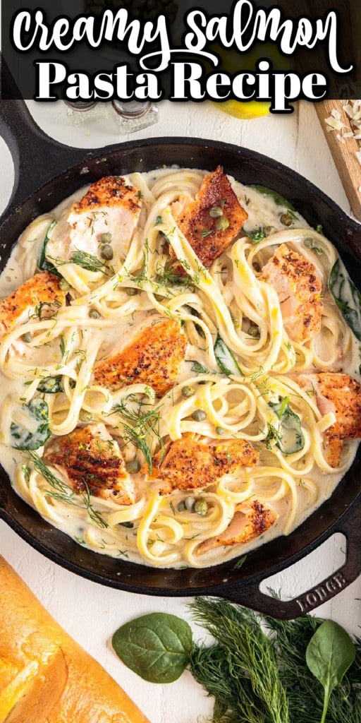 top full view of creamy salmon pasta in skillet next to lemon, capers, salt and pepper, cheese, spices, and bread