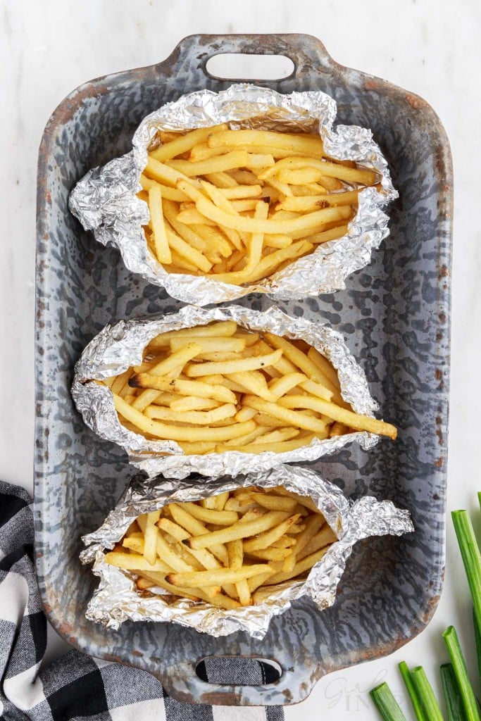 Three foil packets with frozen French fries in a metal baking tray, checkered linen, green onions, on a marble countertop.
