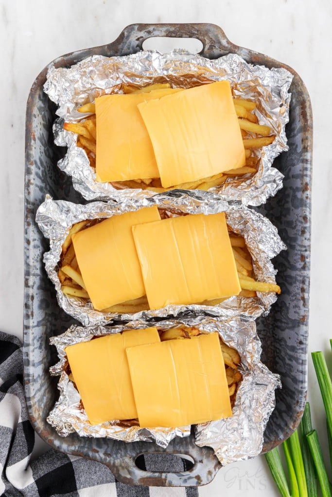 Three foil packets in a baking tray with cooked French fries, topped with sliced cheese, checkered linen, green onions, on a marble countertop.