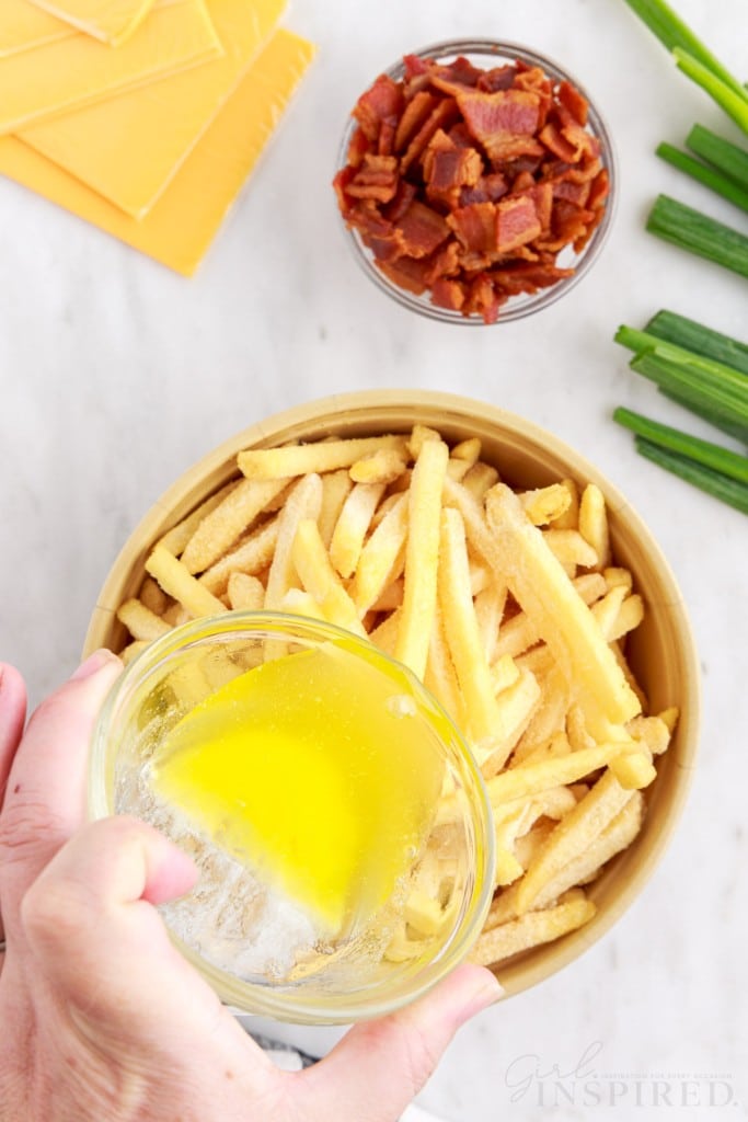 Bowl of frozen French fries, hand pouring melted butter over fries, sliced cheese, small bowl of bacon bits, green onions, on a marble countertop.