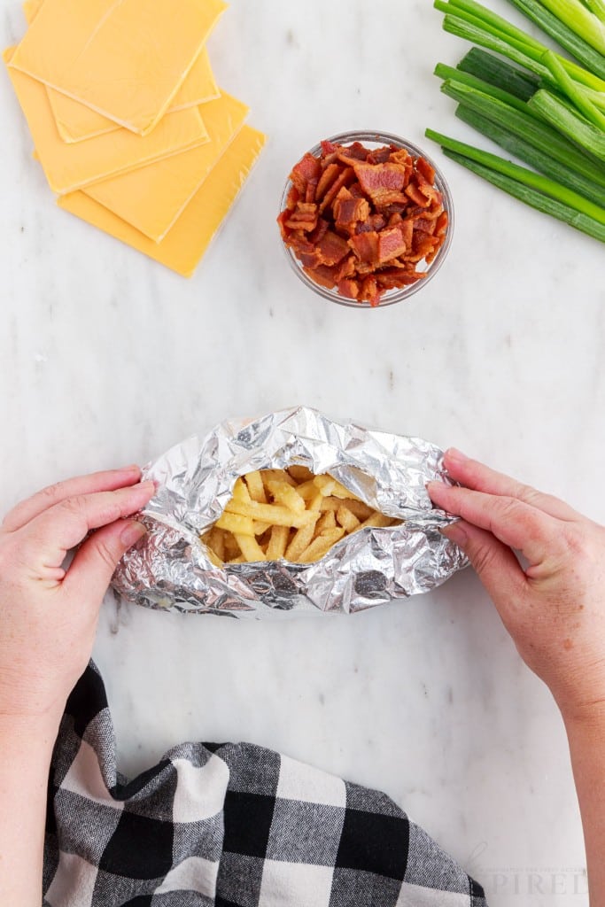 Hands folding foil packet with a small opening at the top, slices of cheese, small bowl of bacon bits, green onions, checkered linen, on a marble countertop.