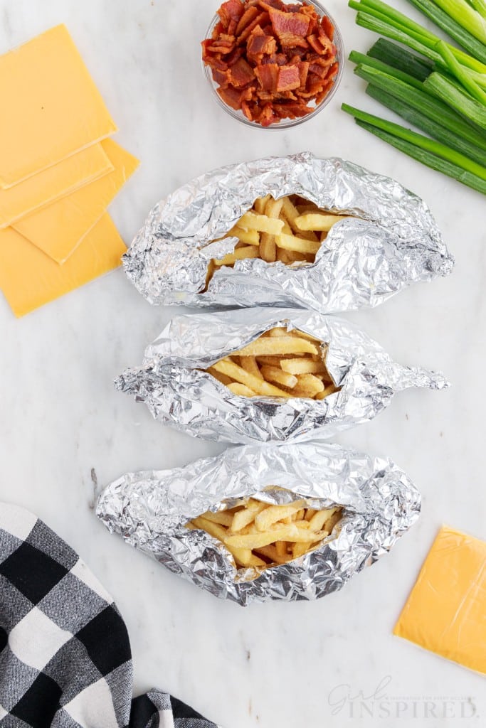 Three foil packets with frozen French fries, slices of cheese, small bowl of bacon bits, green onions, checkered linen, on a marble countertop.