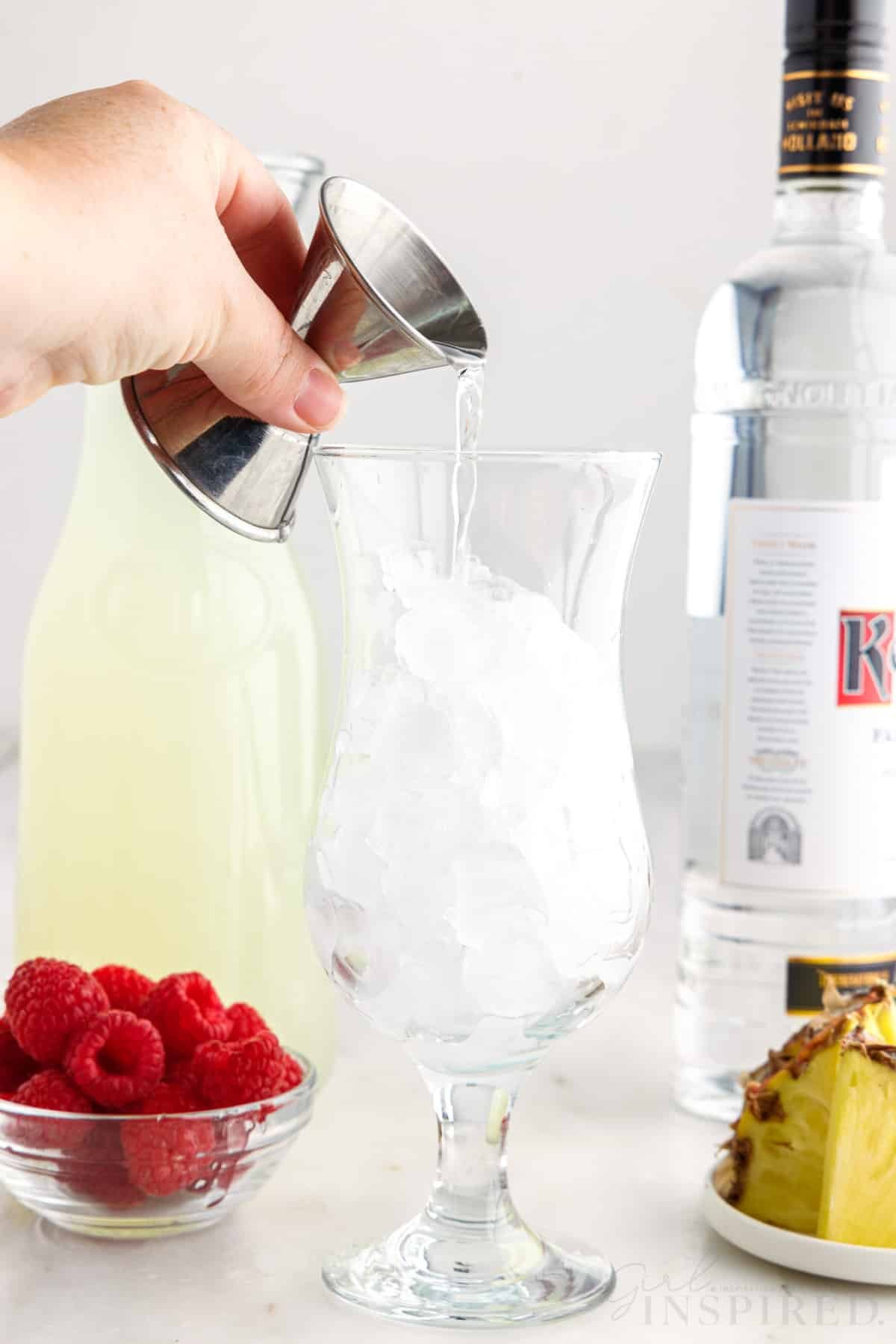 Tall glass filled with ice cubes, hand pouring vodka over the ice, bowl of fresh raspberries, plate of cut pineapple, bottle of vodka, jug of lemonade, on a marble countertop.