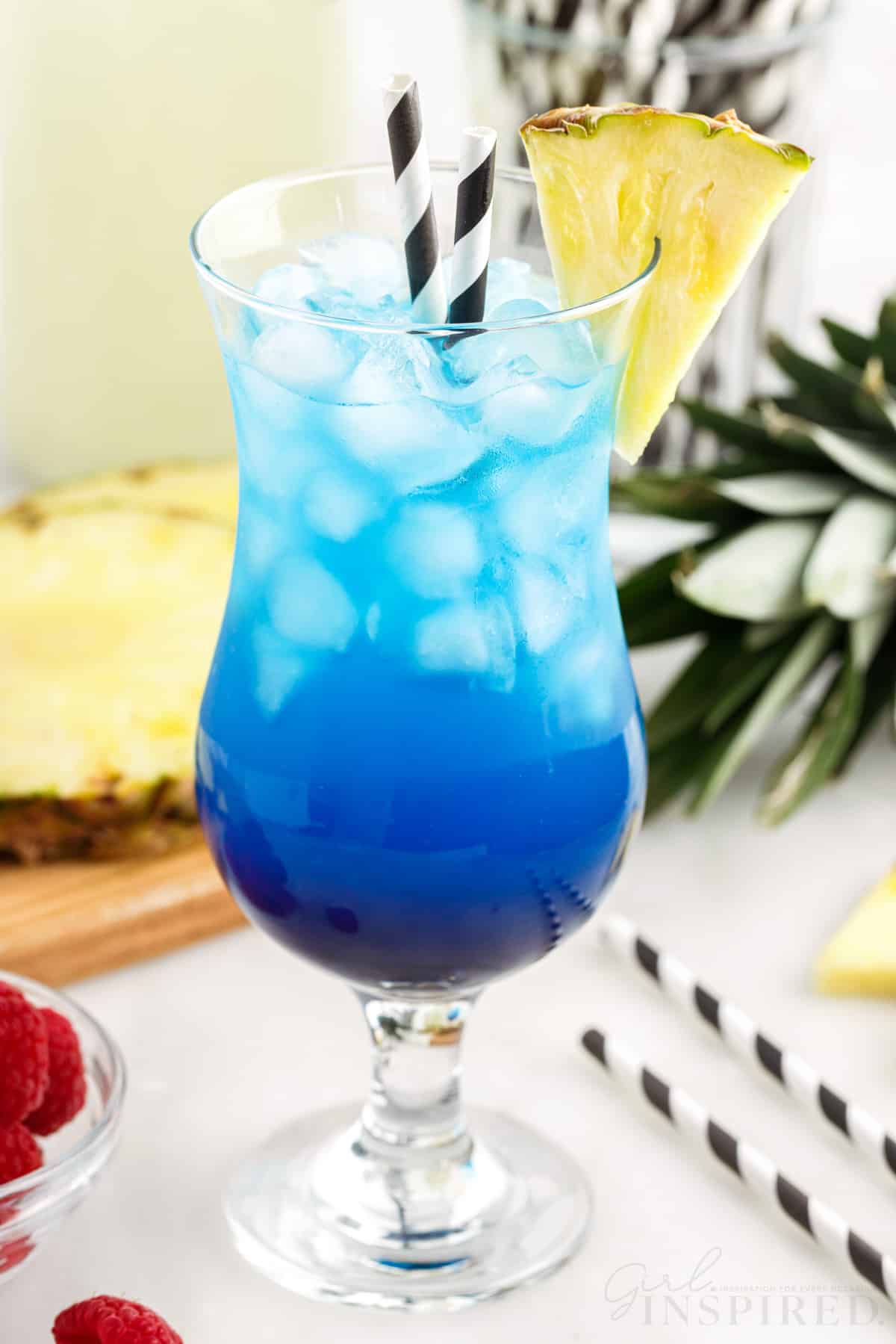 Hurricane style glass with blue fruit tingle cocktail and two black striped straws.